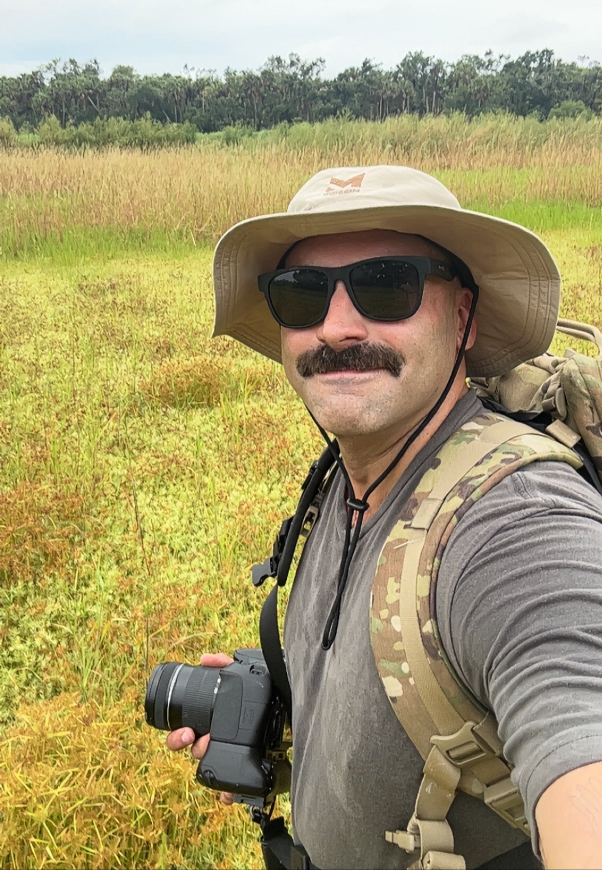 Man in a field wearing a wide-brimmed hat, sunglasses, and a camera, with a backpack.